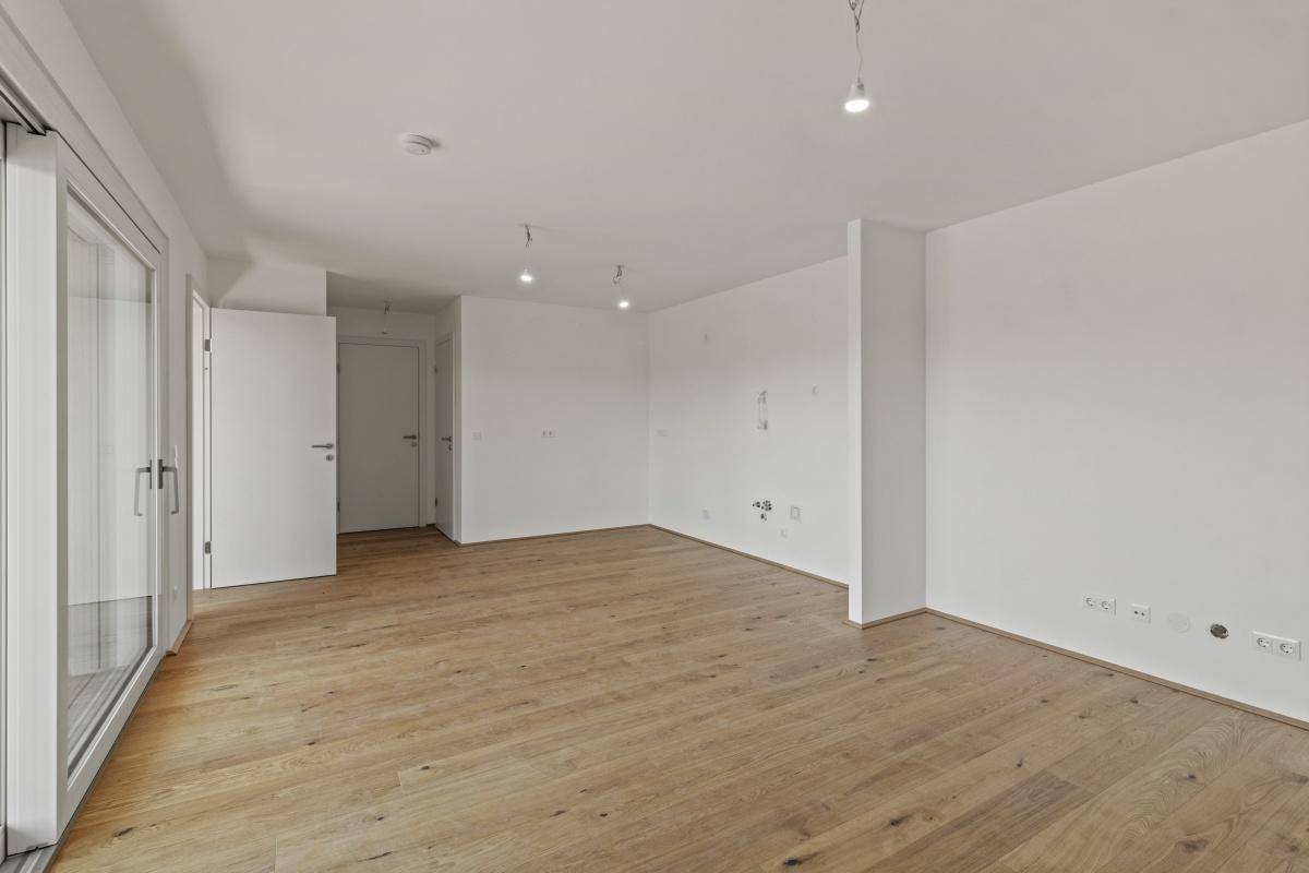 Lichtdurchfluteter, leerer Raum mit Holzboden und breiter Schiebetür zum Balkon.