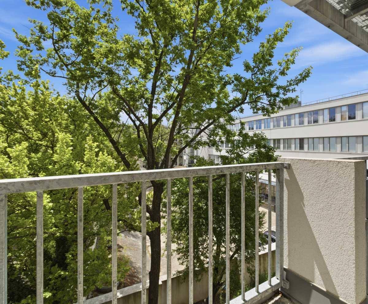 Blick vom Balkon ins Grüne mit Bäumen und einem Gebäude im Hintergrund.