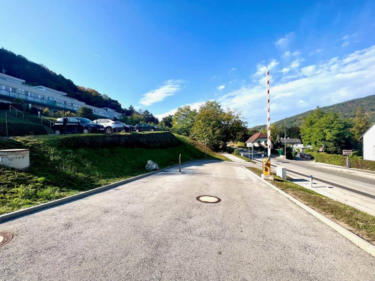 Asphaltierter Parkplatz mit Schranke und Zufahrt zu einer Straße, umgeben von grünen Hügeln.