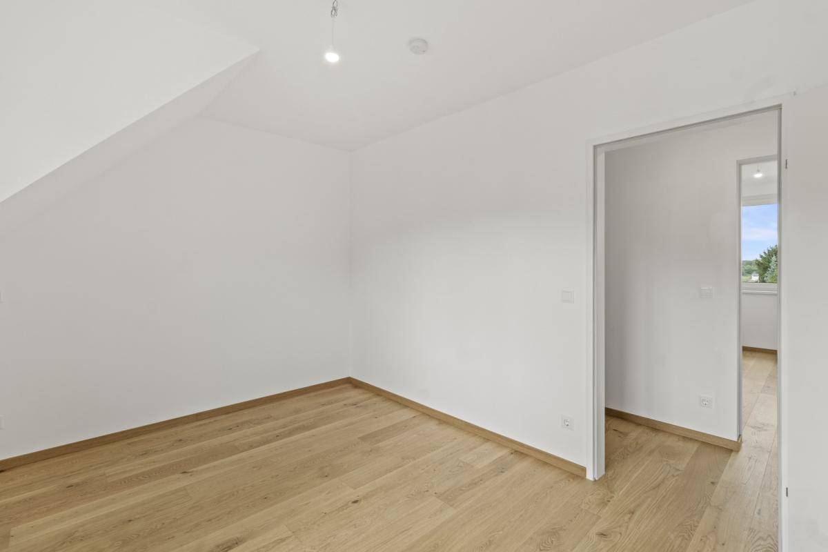 Heller Raum mit Holzboden, weißen Wänden und Dachschräge, offen zu einem weiteren Bereich.