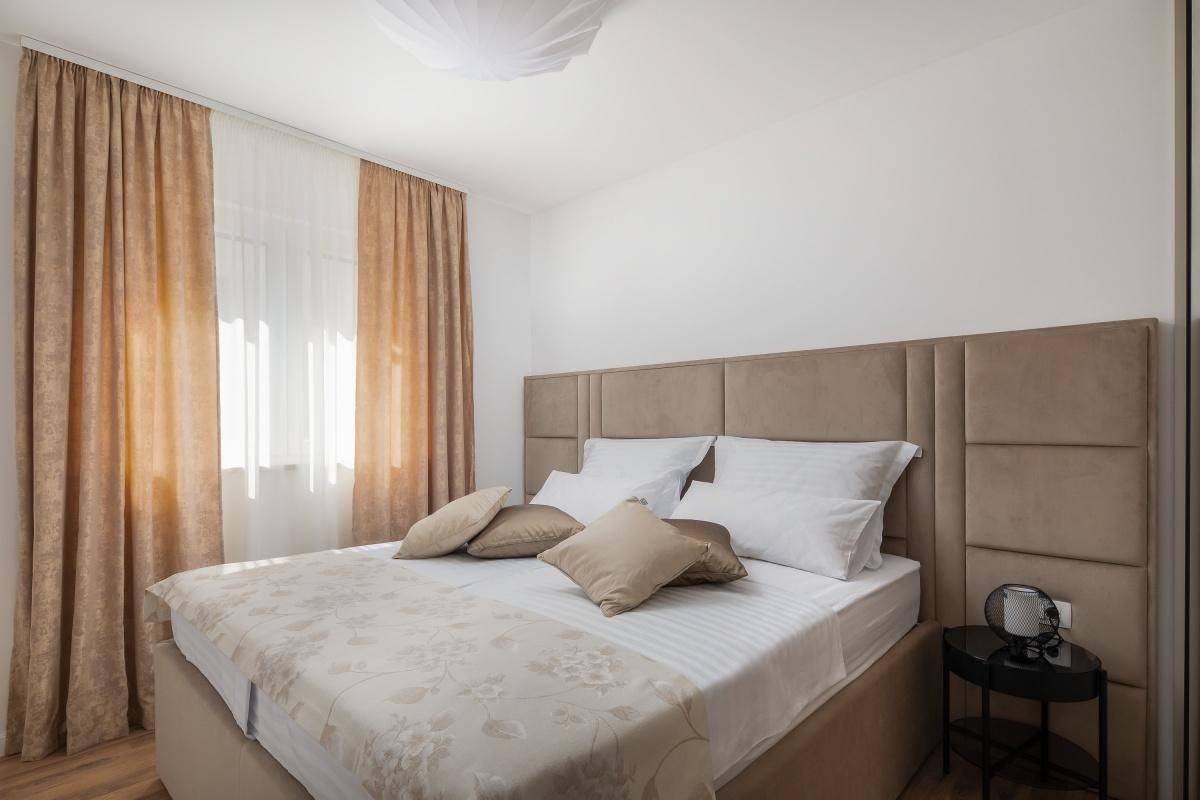 Gemütliches Schlafzimmer mit großem Doppelbett, beigem Kopfteil und Vorhängen, sowie Holzboden.