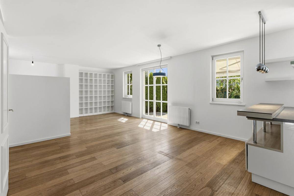 Helles, leeres Wohnzimmer mit Holzboden, großen Fenstern und Blick ins Grüne.