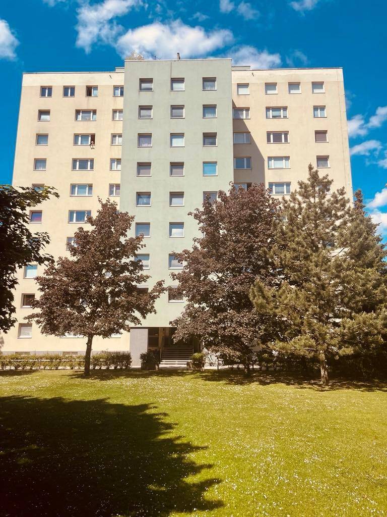 Mehrstöckiges Wohngebäude mit heller Fassade und grünen Akzenten, umgeben von Bäumen und Rasenflächen.