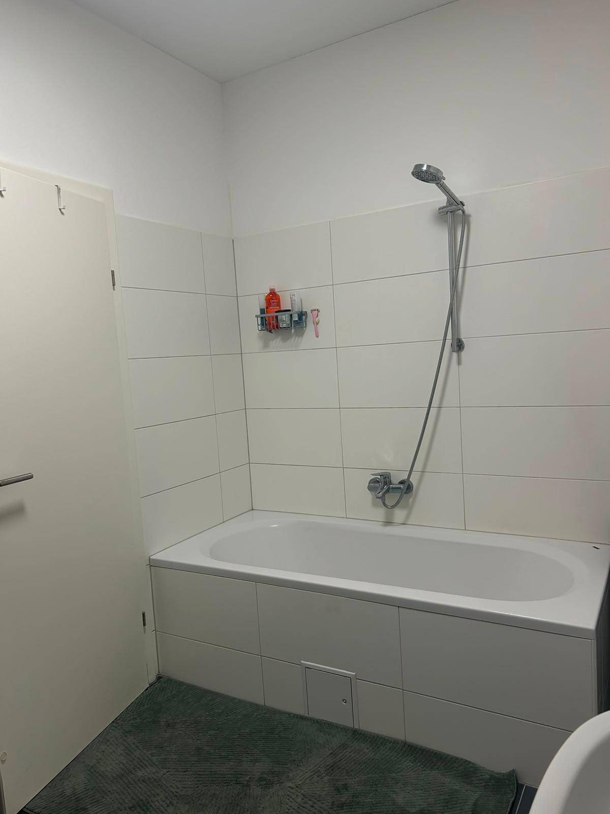 Badezimmer mit Badewanne und Duschvorrichtung, umgeben von weißen Fliesen.
