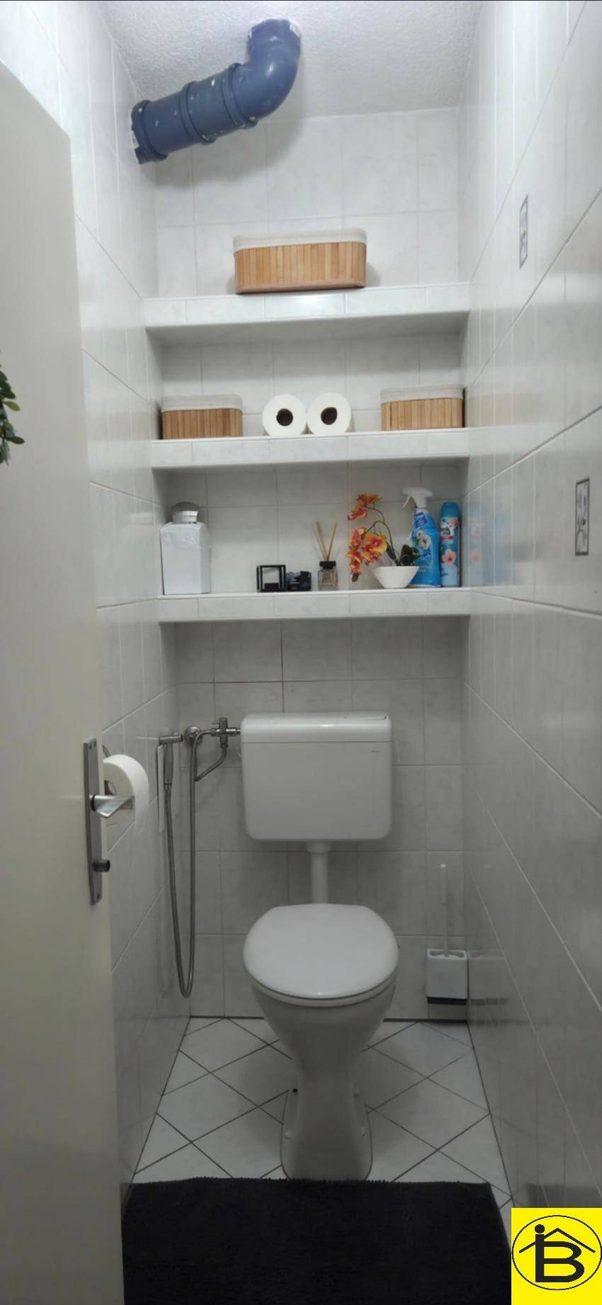Separates WC mit Toilette, weißen Fliesen, praktischen Wandregalen und Abluftrohr.