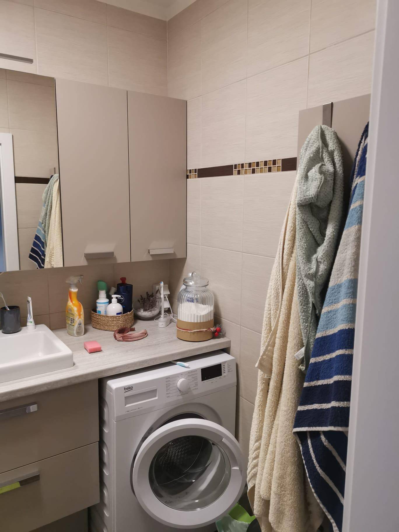 Badezimmer mit Waschmaschine, Waschbecken, Spiegelschrank und beigen Fliesen.