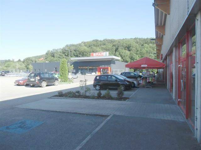 Außenansicht eines Supermarktes mit großem Parkplatz und bewaldetem Hügel im Hintergrund.
