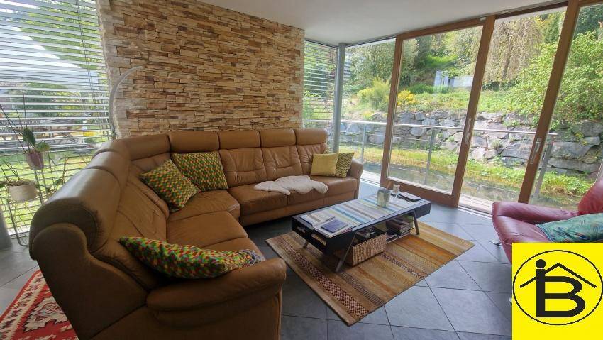 Helles Wohnzimmer mit bequemer Ledercouch, Steinwand und großen Fenstern mit Blick ins Grüne.