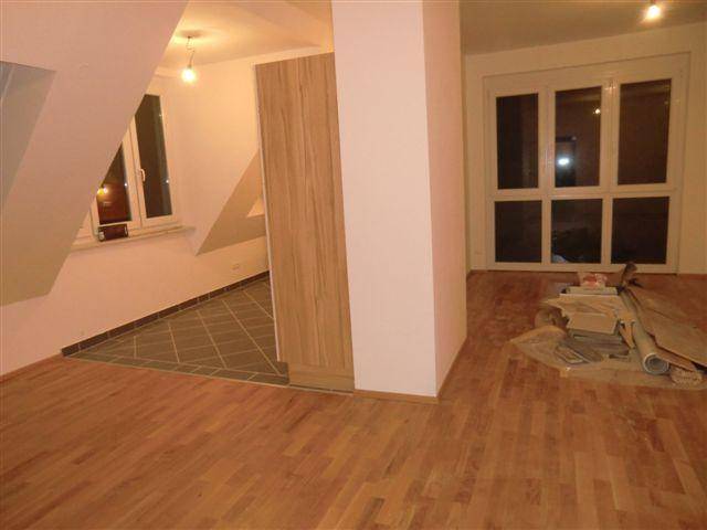 Geräumiger Wohnbereich mit Holzboden, großen Fenstern und Zugang zu einem weiteren Raum.