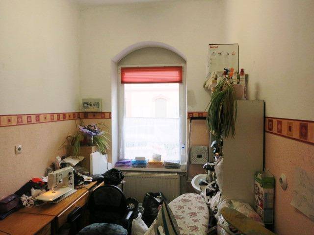 Arbeitsraum mit Nähmaschine, einem Bogenfenster mit rotem Rollo und verschiedenen Arbeitsmaterialien, hell gestrichene Wände.