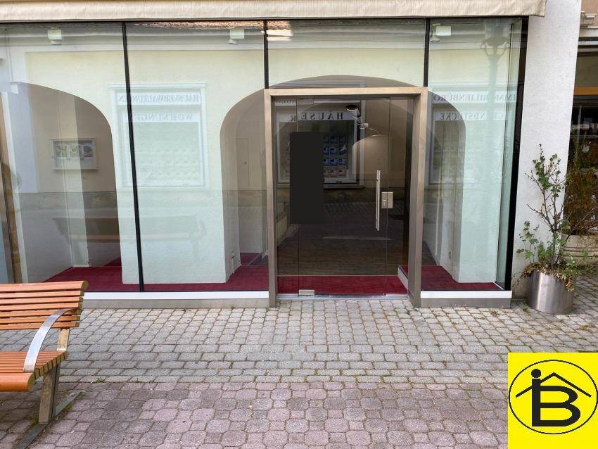 Moderne Ladenfront mit großer Glasfläche und gläserner Eingangstür, davor ein gepflasterter Bereich.