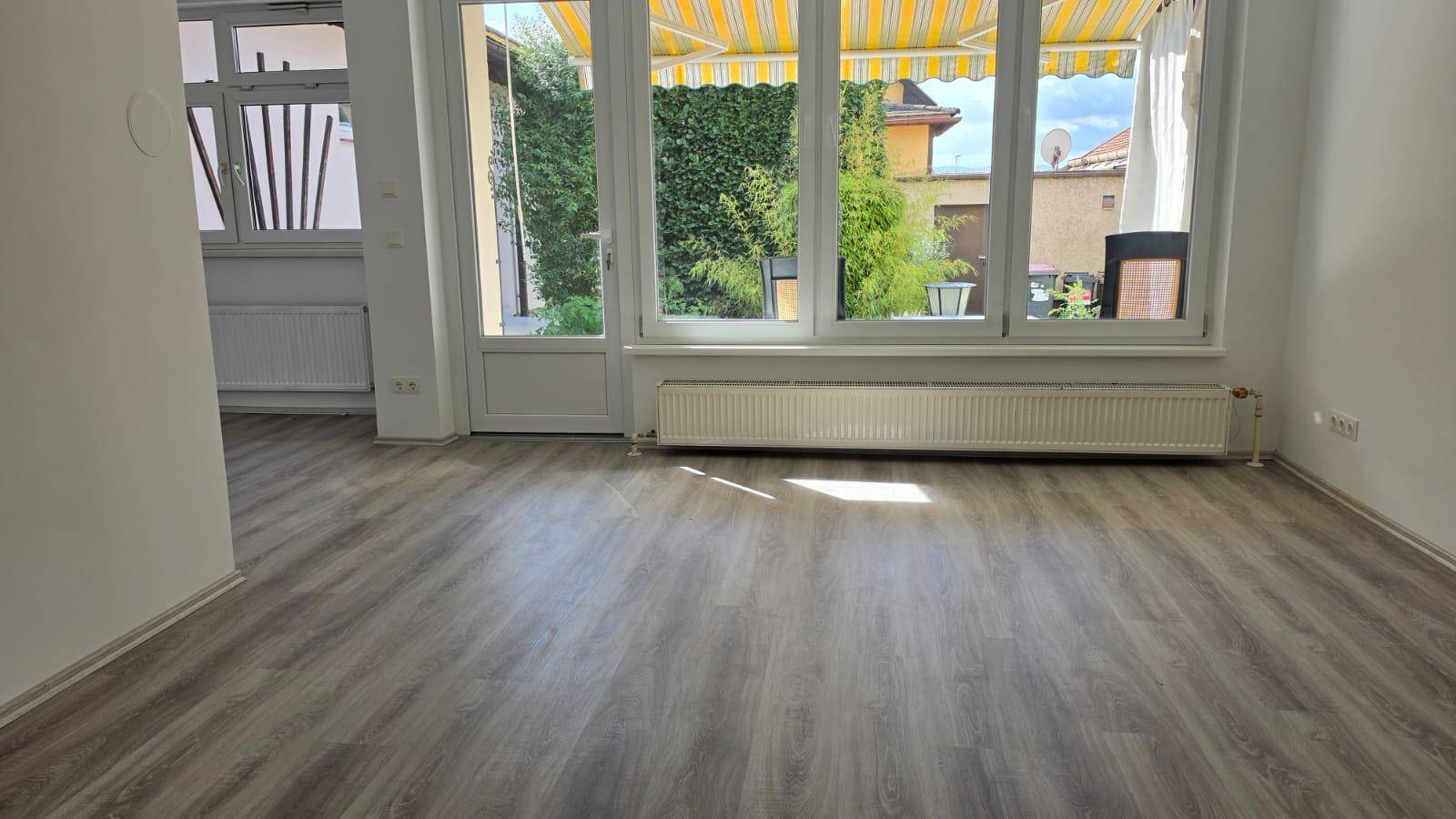 Lichtdurchfluteter Wohnbereich mit Laminatboden und großen Fenstern zum grünen Außenbereich.