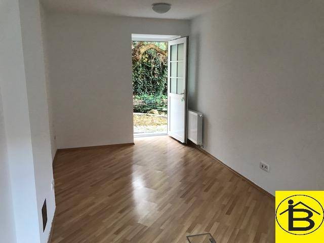 Leerer Raum mit Holzparkettboden und einer Tür, die direkt in einen begrünten Außenbereich führt.