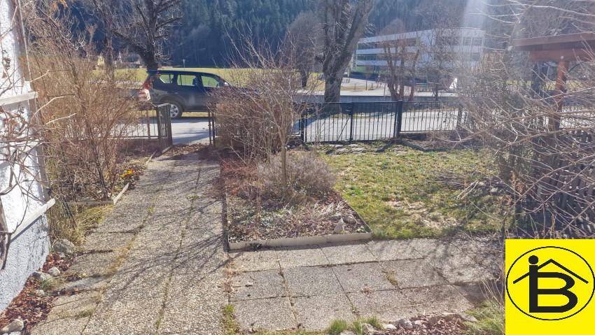 Vorgarten mit gepflastertem Weg und Rasenfläche, umgeben von Sträuchern und Bäumen.