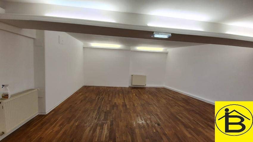 Leerer Raum mit Holzboden, weißen Wänden und niedriger Decke, ausgestattet mit Heizkörpern.