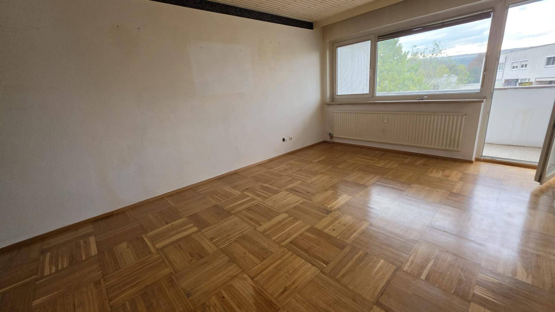 Heller Raum mit Fischgrätparkettboden, großem Fenster und Zugang zu einem Balkon.