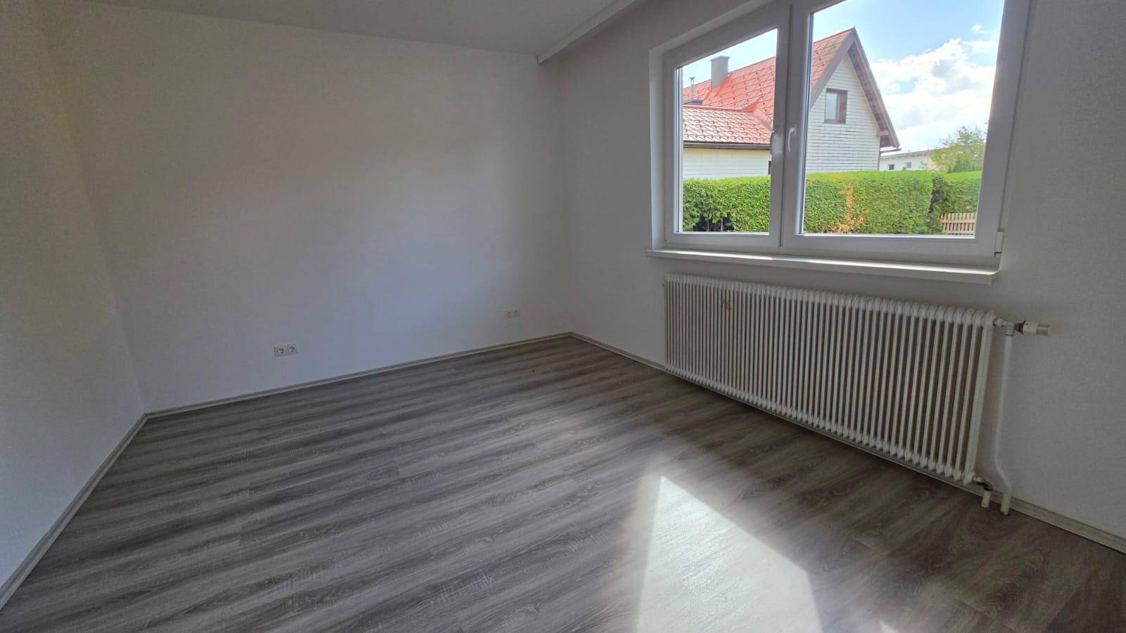 Sonniger Raum mit Laminatboden und großem Fenster mit Blick auf ein Nachbarhaus und Grünflächen.