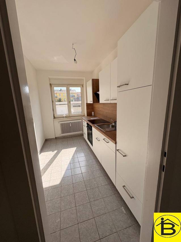 Helle Küche mit Fenster, weißen Schränken und Fliesenboden, bietet ausreichend Platz und Tageslicht.