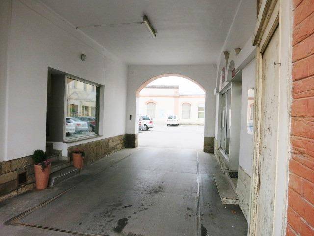 Überdachter Durchgang mit einem Bogen, der zu einem offenen Bereich mit parkenden Autos führt, weiße Wände und Betonboden.