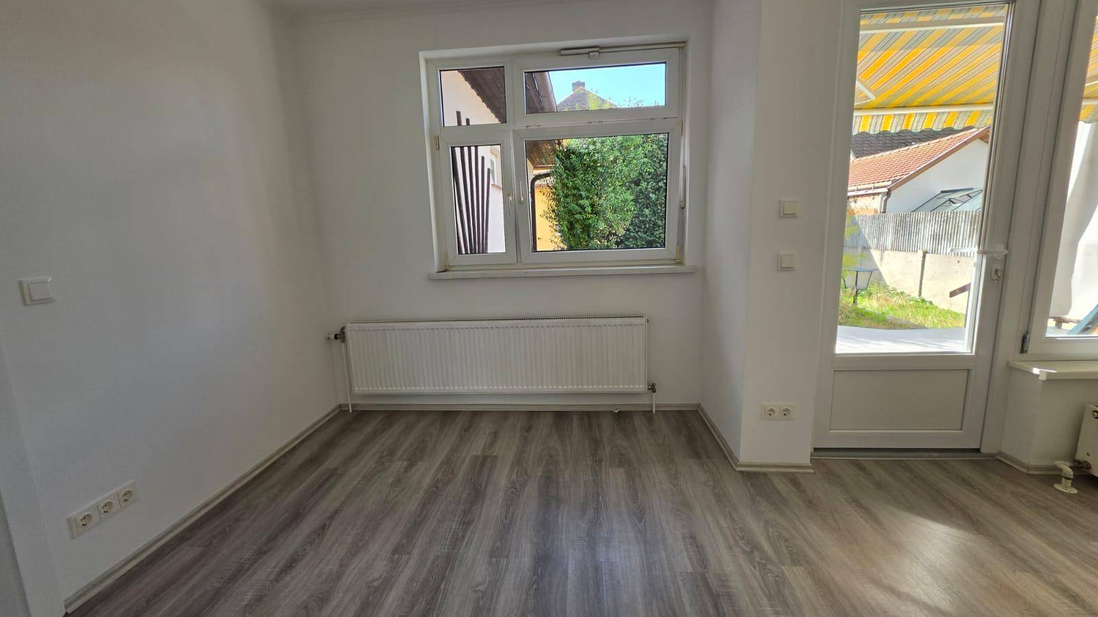 Heller Raum mit Laminatboden, großem Fenster und Zugang zu einem Außenbereich mit Markise.