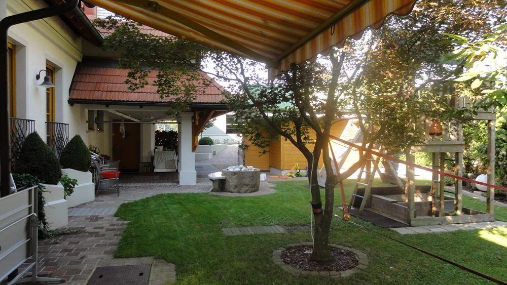 Gartenbereich mit überdachter Terrasse, einem Baum und einem Spielplatz für Kinder.
