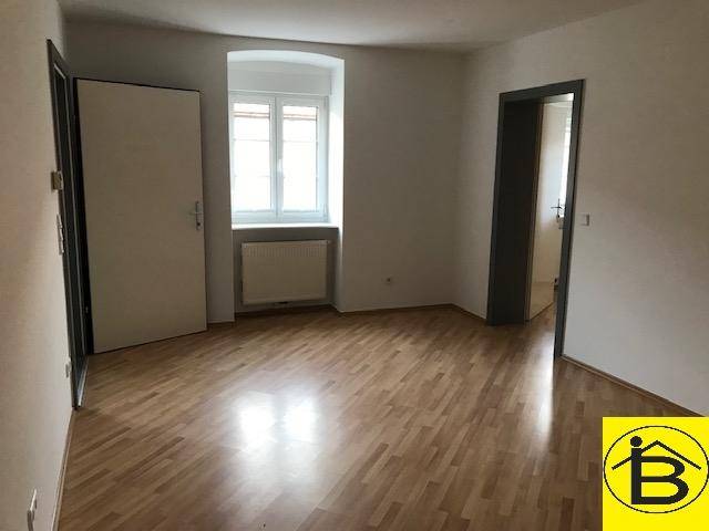 Leerer Raum mit Holzparkettboden, weiß gestrichenen Wänden und einem Fenster mit Heizkörper.