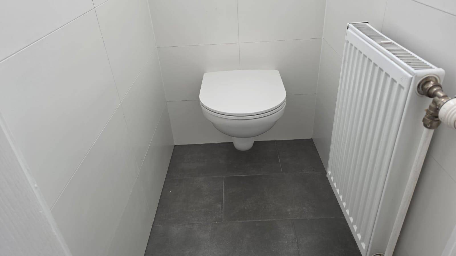 Separates WC mit wandhängender Toilette, weißen Fliesen und Heizkörper.
