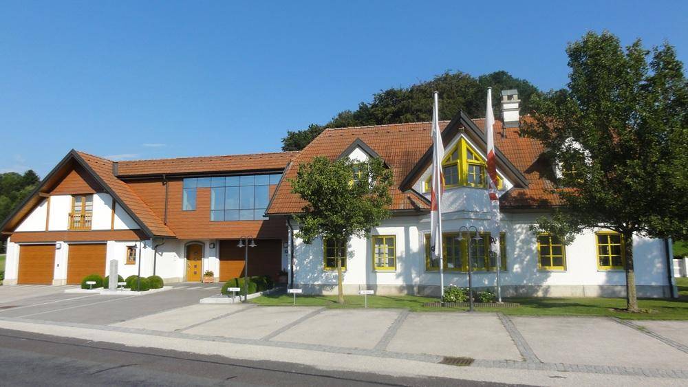 Hauptgebäude mit gelben Fensterrahmen, Fahnenmasten und einem modernen Glasanbau.