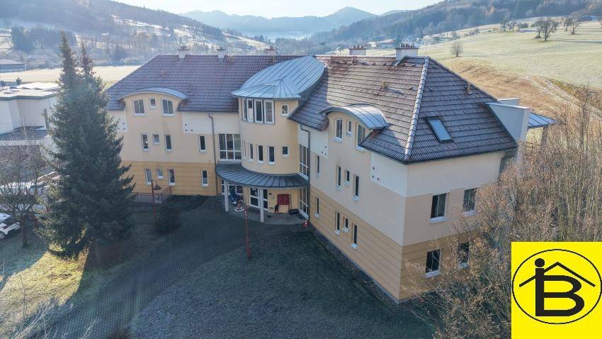 Vogelperspektive des Mehrfamilienhauses mit gelber Fassade, rotem Dach und umliegender Landschaft.
