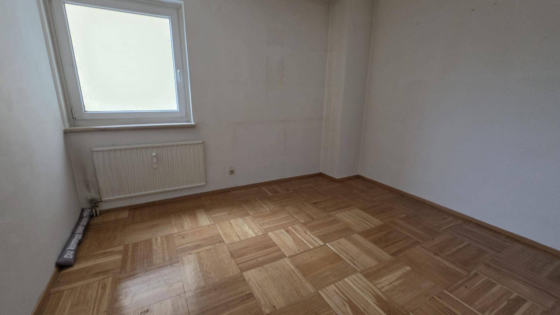 Heller Raum mit klassischem Parkettboden und einem Fenster, das Tageslicht hereinlässt.