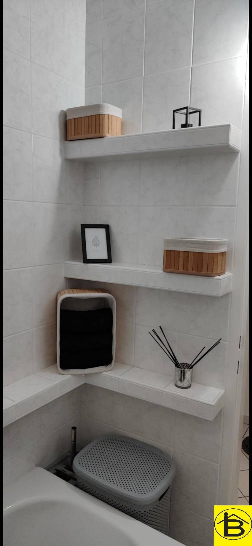 Badezimmer mit weißen Fliesen, schwebenden Regalen und dem Rand einer Badewanne.