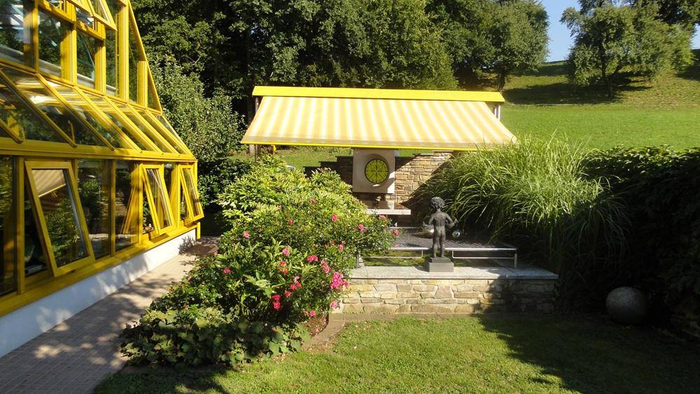 Gartenbereich mit Glas-Wintergarten, Markise und einer kleinen Statue in der Grünanlage.