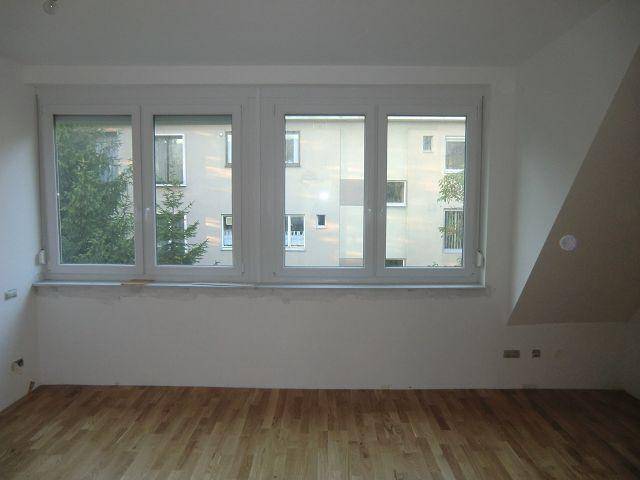 Helles Zimmer mit Holzboden und großen Fenstern, die den Blick auf Gebäude freigeben.