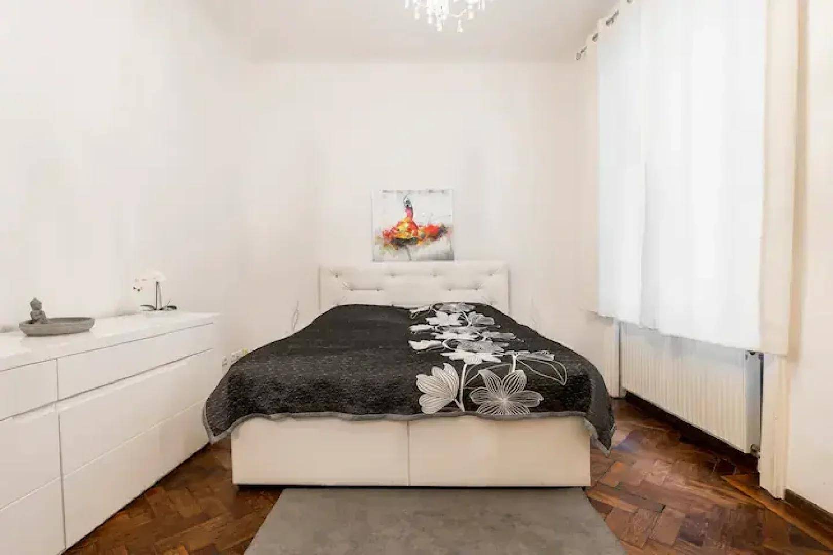 Schlafzimmer mit Doppelbett, weißer Kommode und Parkettboden, beleuchtet durch natürliches Licht.