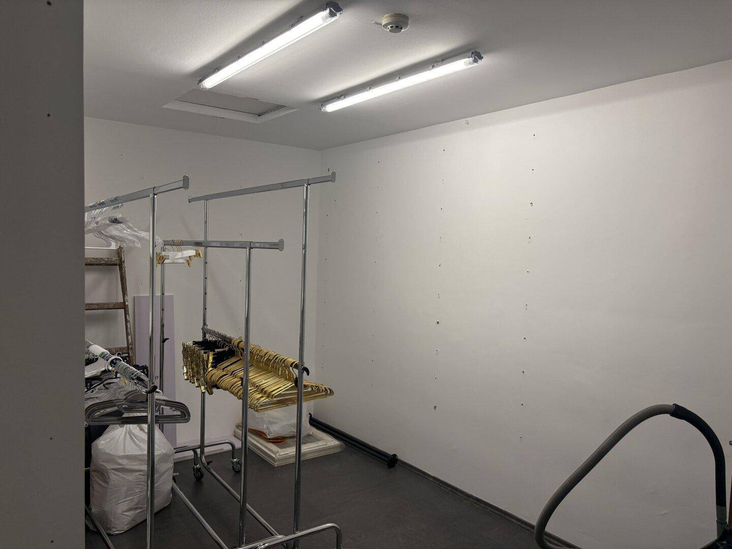 Abstellraum oder Lagerbereich mit Kleiderständern, dunklem Boden und Deckenbeleuchtung.