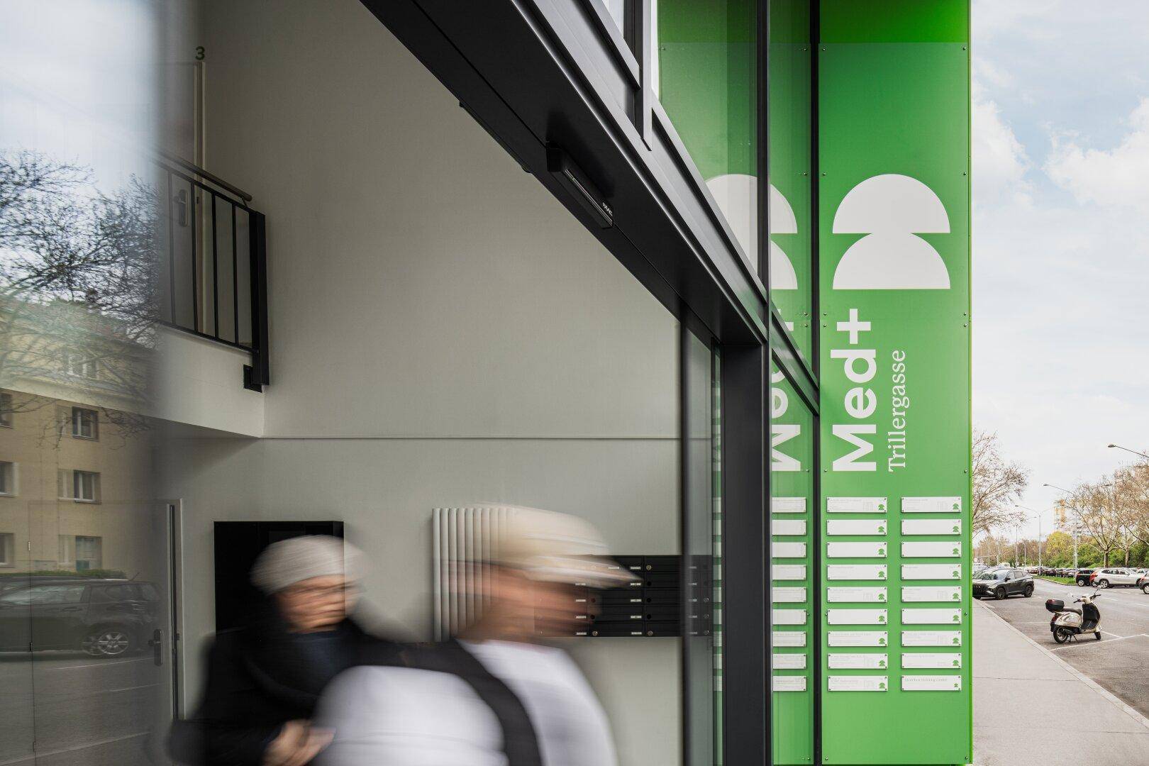 Eingangsbereich eines modernen Gebäudes mit grüner Beschilderung und verglasten Türen, Personen betreten das Gebäude.