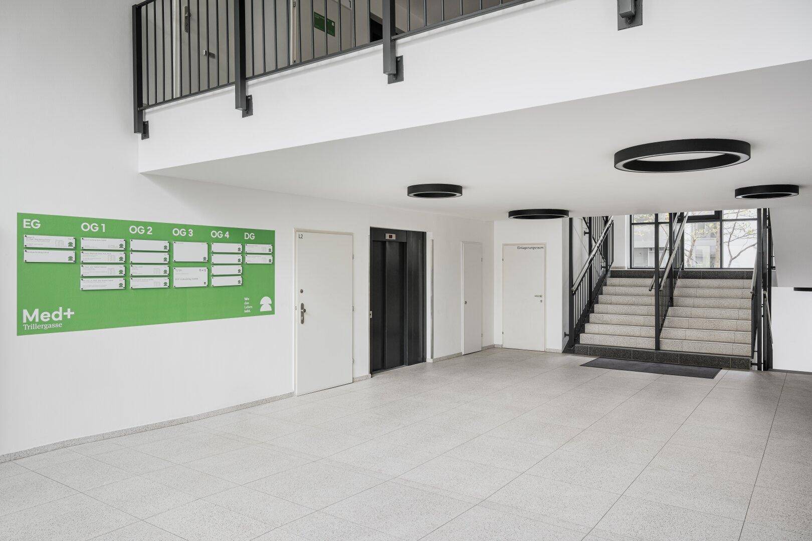 Heller und geräumiger Eingangsbereich mit Fliesenboden, Aufzug, Treppe und einem großen Informationsschild.