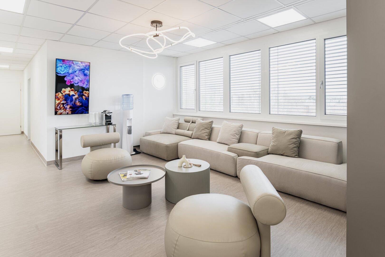 Komfortabler Wartebereich mit bequemen Sofas, modernen Tischen und großen Fenstern, die viel Tageslicht hereinlassen.