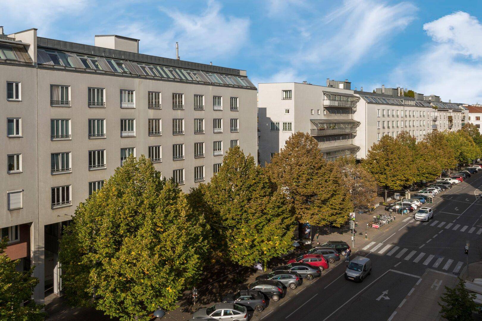 Straßenansicht von Wohngebäuden mit parkenden Autos und Bäumen an einem sonnigen Tag.