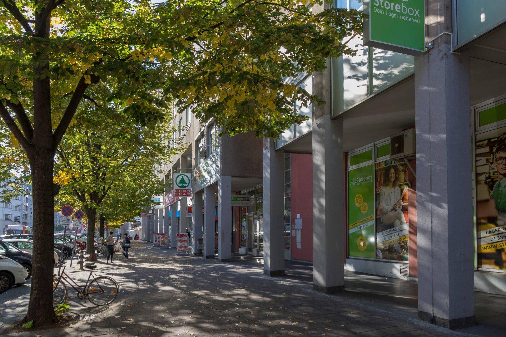 Erdgeschoss eines Wohngebäudes mit Geschäften, Bürgersteig und Bäumen an einem sonnigen Tag.
