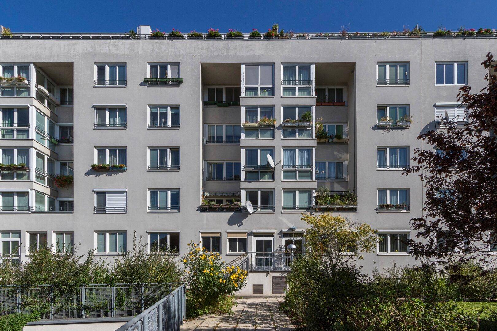 Fassade eines Wohngebäudes mit zahlreichen Fenstern, Balkonen und einer kleinen Grünfläche davor.
