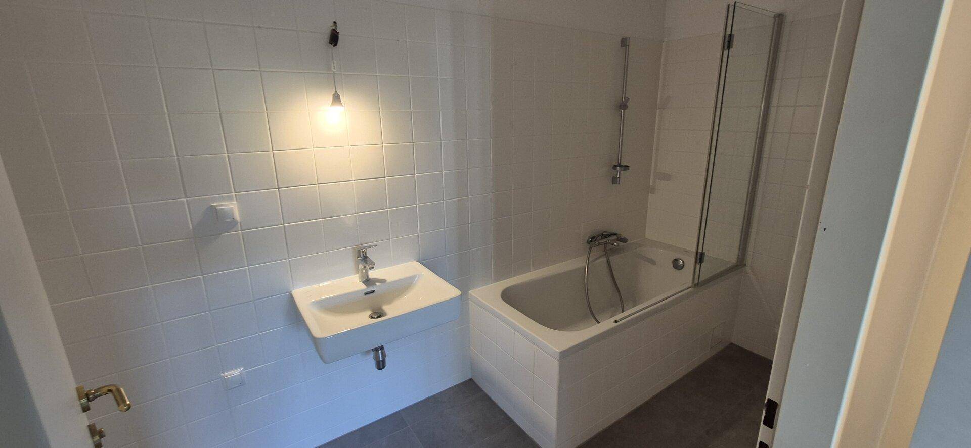 Badezimmer mit weißen Fliesen, Badewanne mit Duschwand und einem einzelnen Waschbecken.