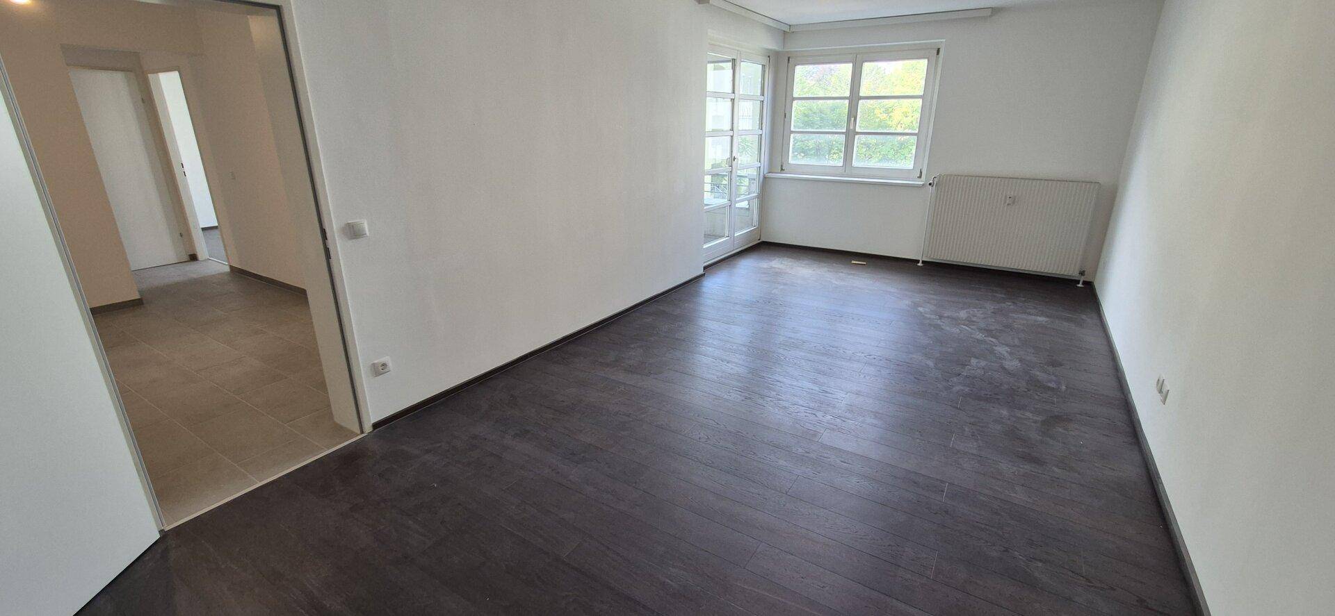 Leeres Zimmer mit dunklem Boden, weißen Wänden, Fenster und Heizkörper, bereit zur Einrichtung.