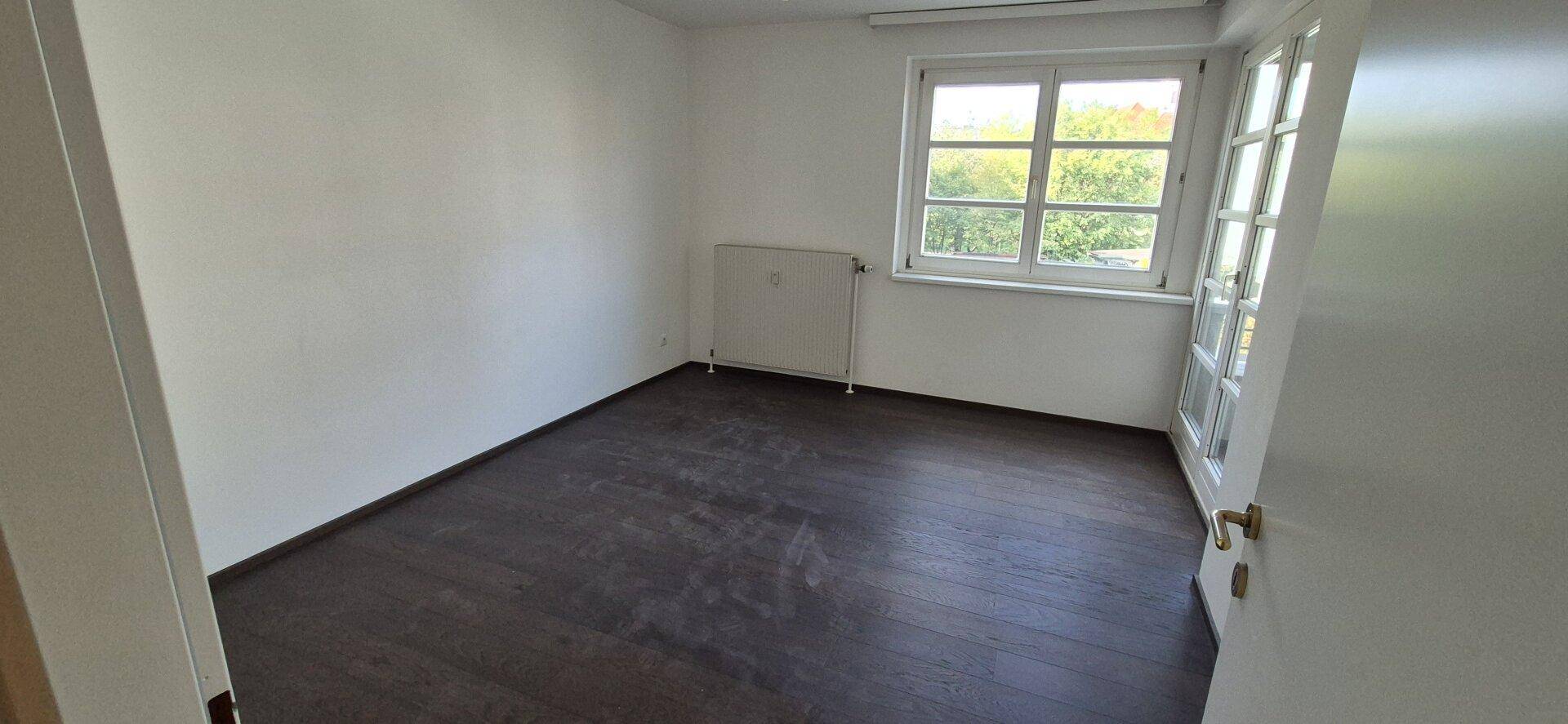 Helles, leeres Zimmer mit dunklem Boden, weißen Wänden und großem Fenster.