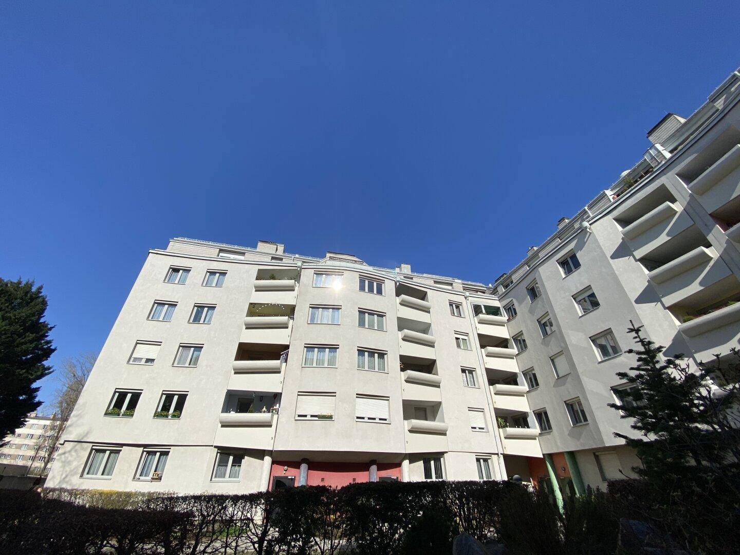 Mehrstöckiges Wohngebäude mit zahlreichen Balkonen und Fenstern unter strahlend blauem Himmel.