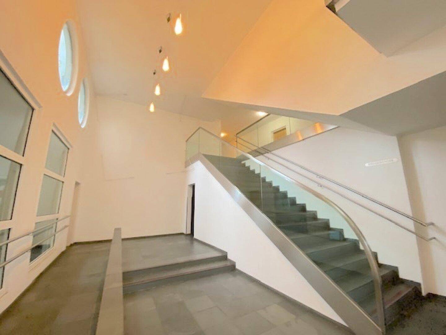 Moderner Eingangsbereich mit eleganter Treppe, Glasgeländer und runden Fenstern, die viel Licht spenden.