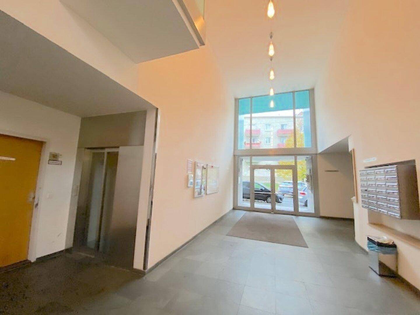 Heller Eingangsbereich mit Aufzug, Briefkästen und großen Glasfenstern zur Straße hin.
