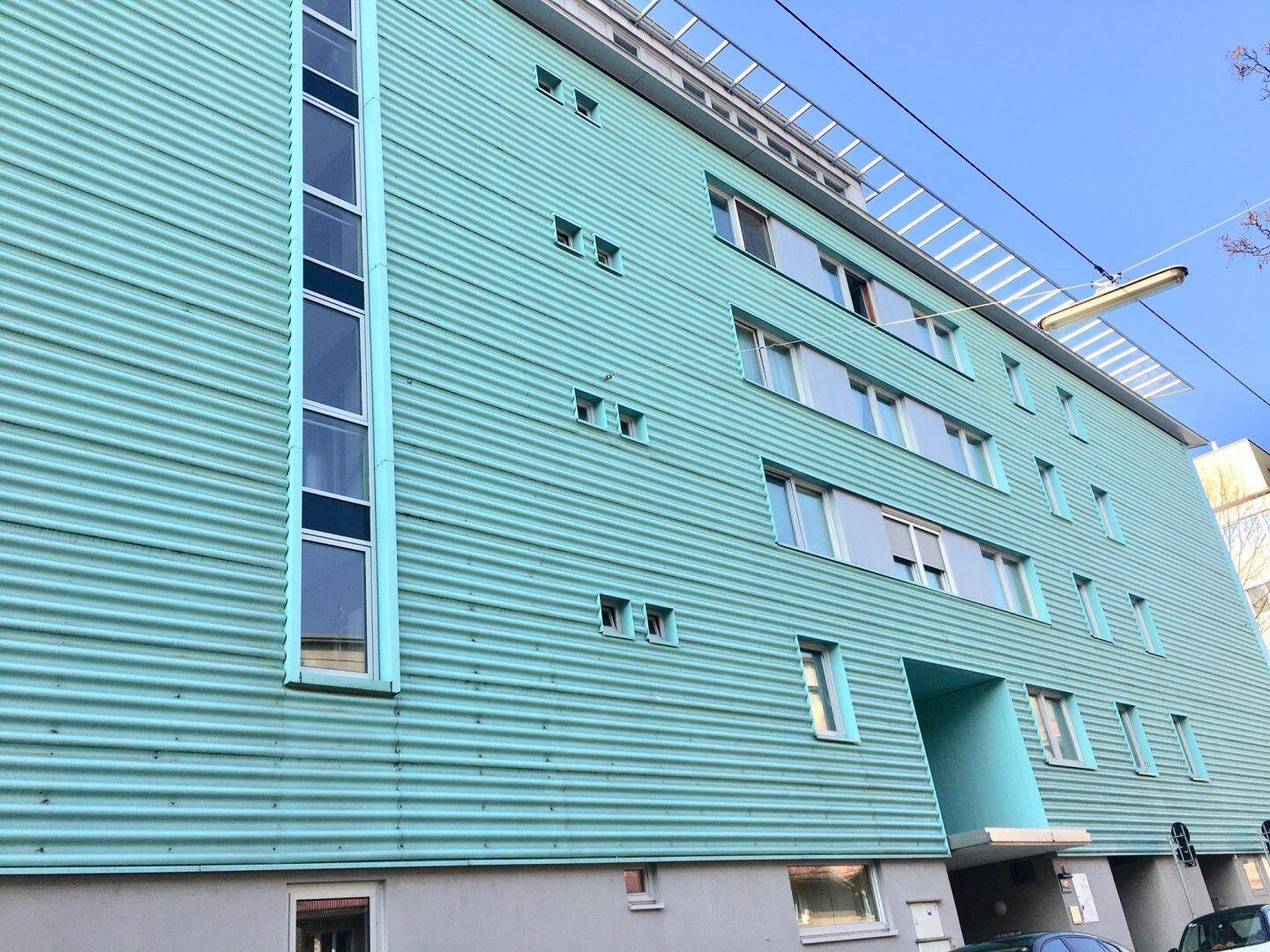 Moderne Gebäudefassade in leuchtendem Türkis mit großen Fenstern und klaren Linien.