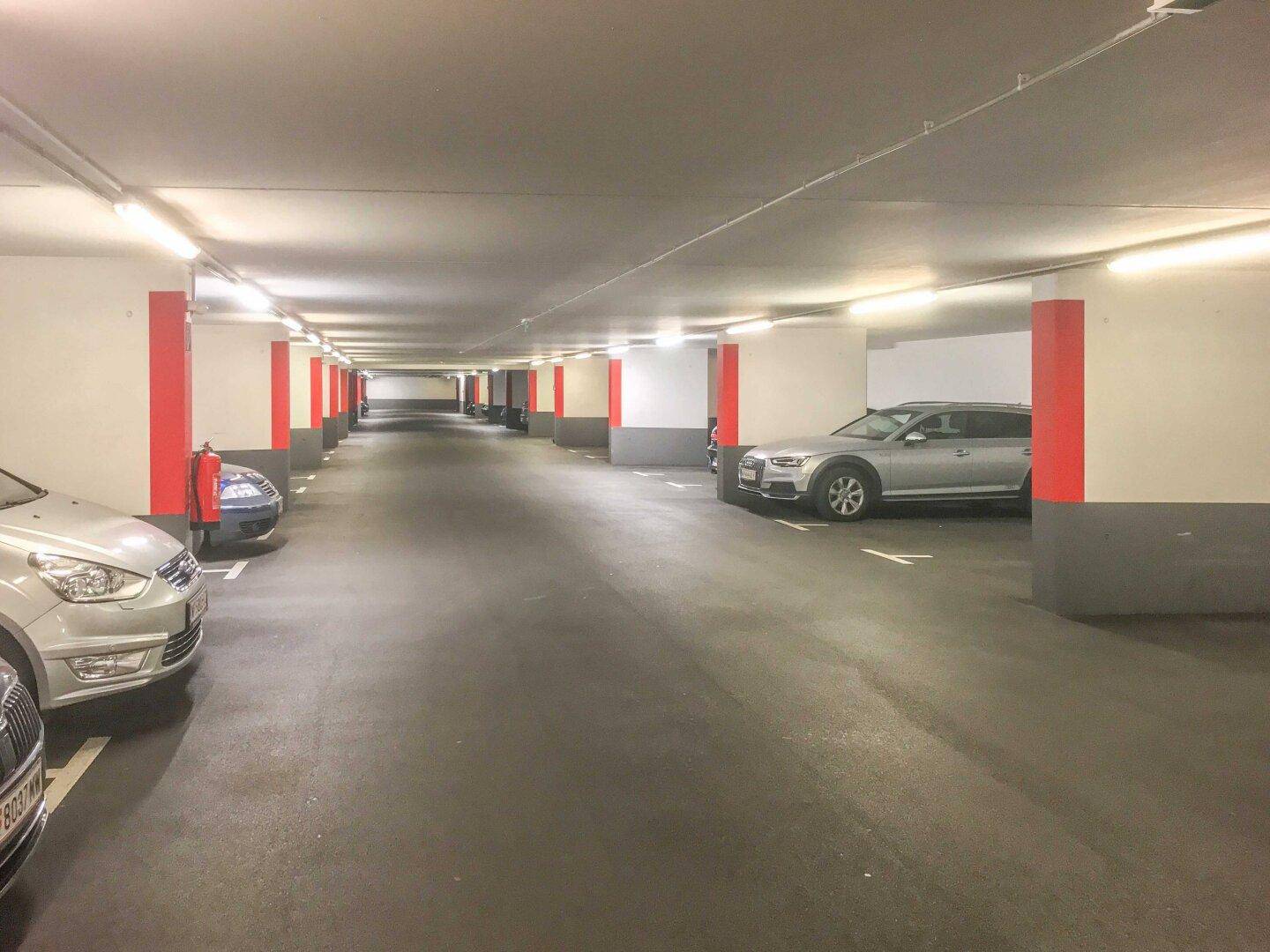 Weitläufige Tiefgarage mit zahlreichen Stellplätzen, roten Säulen und heller Deckenbeleuchtung.