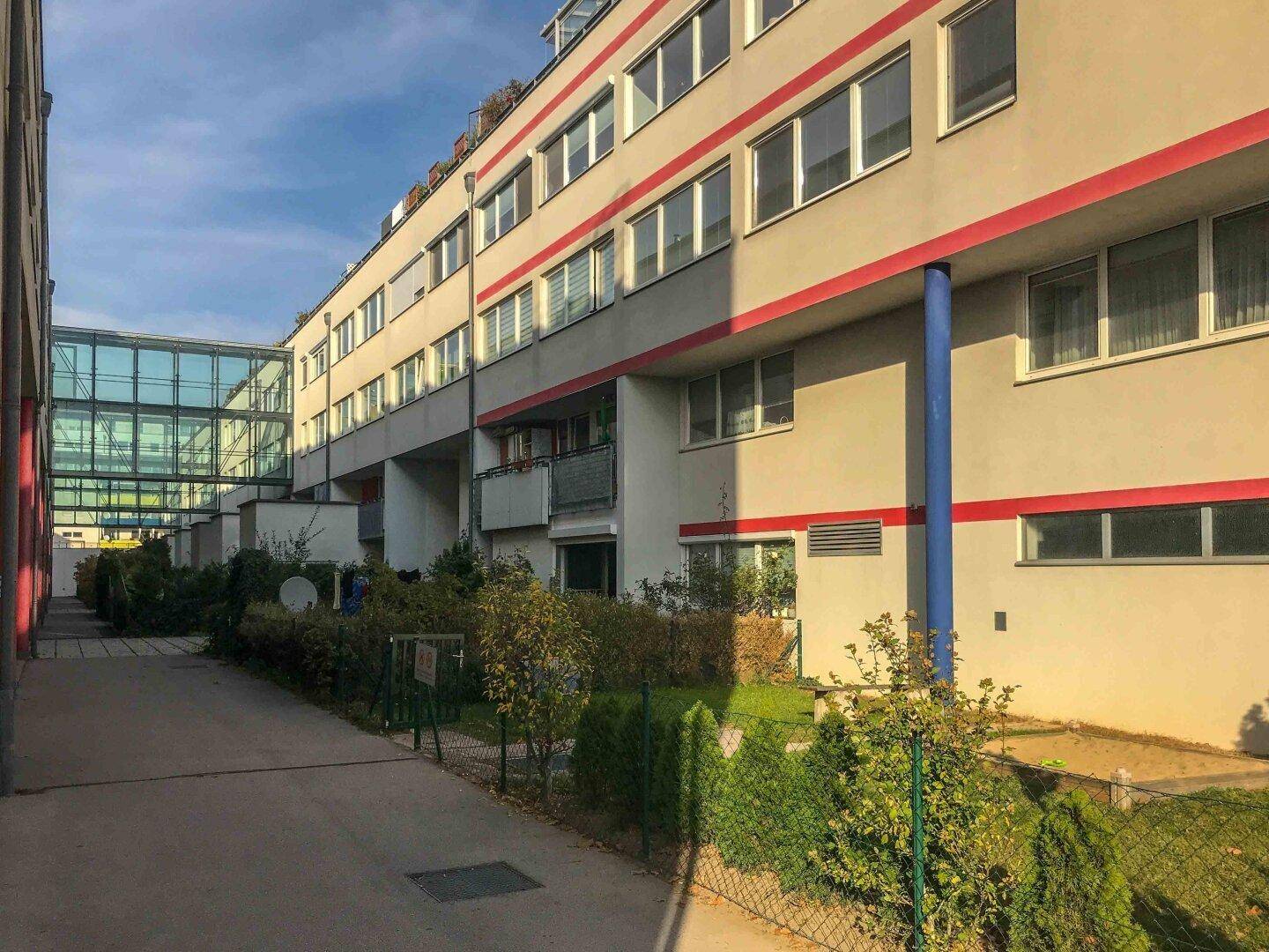 Außenansicht eines Wohnkomplexes mit langen Balkonen, Grünflächen und einem verglasten Übergang zwischen Gebäudeteilen.